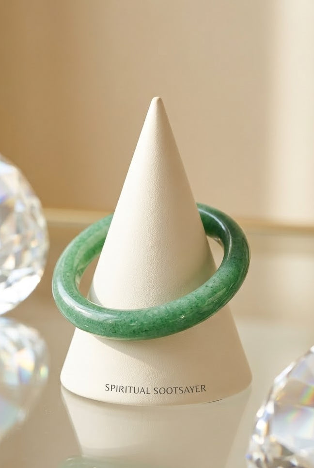 A green jade bangle bracelet displayed on a white cone-shaped jewelry stand, with faceted crystal props on both sides against a warm beige background.