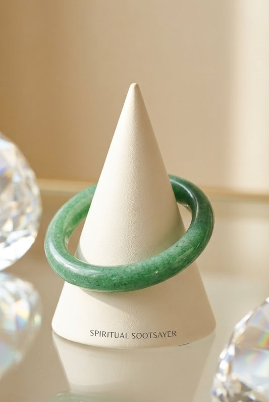 A green jade bangle bracelet displayed on a white cone-shaped jewelry stand, with faceted crystal props on both sides against a warm beige background.