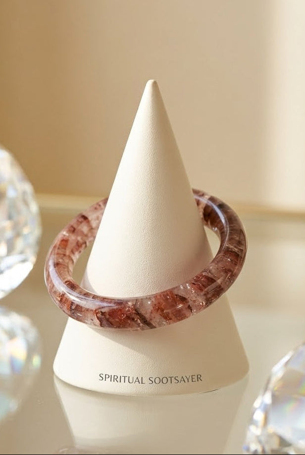 A polished reddish-brown crystal bangle bracelet displayed on a white cone stand labeled 'SPIRITUAL SOOTSAVER', with blurred faceted crystal spheres in the background.