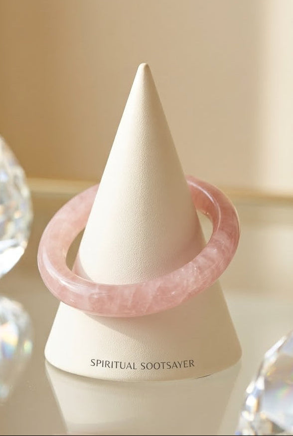A light pink stone bangle bracelet displayed on a white cone stand, with the brand name 'SPIRITUAL SOOTSAVER' printed on the base. The stand is set on a reflective surface with decorative clear crystal spheres in the background.