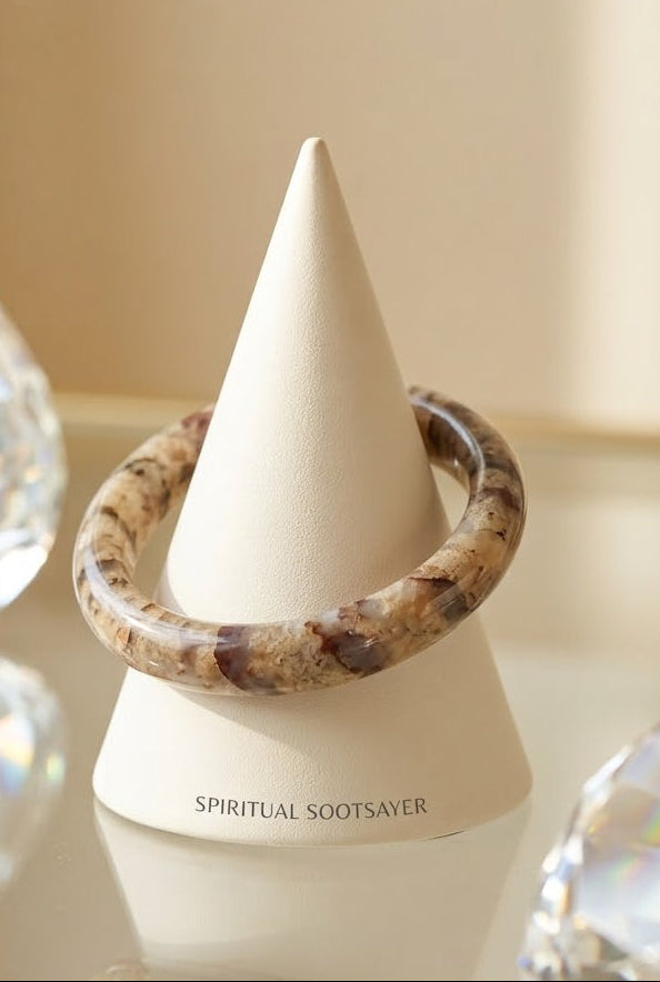 A brown and beige marbled bangle bracelet displayed on a white cone stand labeled 'SPIRITUAL SOOTSAVER', set on a reflective surface with blurred crystal props.