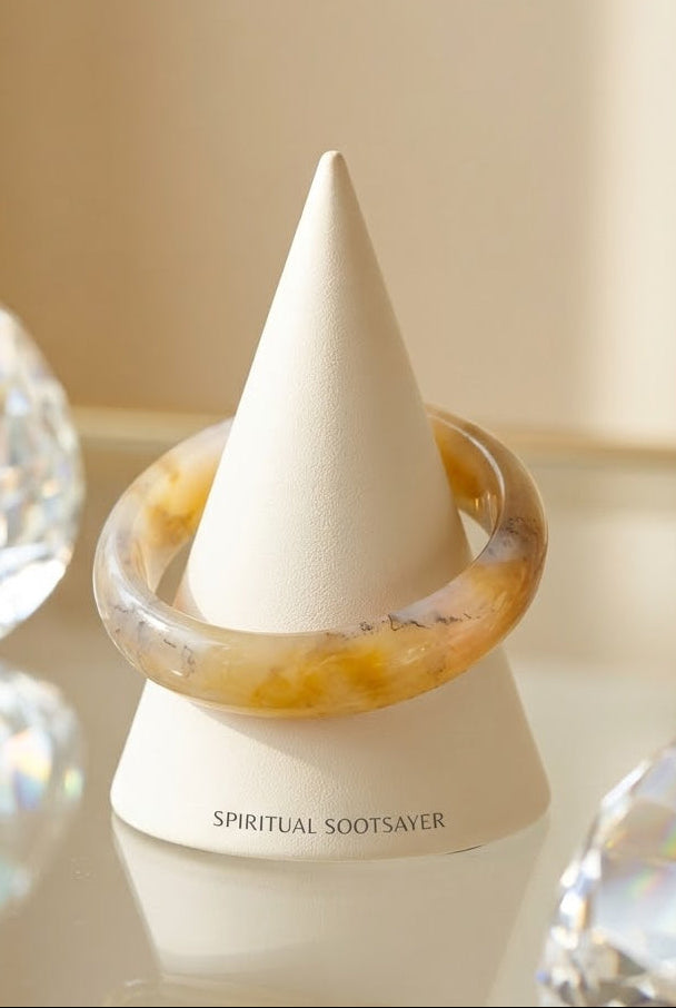 A translucent amber-yellow bangle bracelet displayed on a white cone stand labeled 'SPIRITUAL SOOTSAVER', with blurred faceted crystal-like props on both sides against a warm beige background.