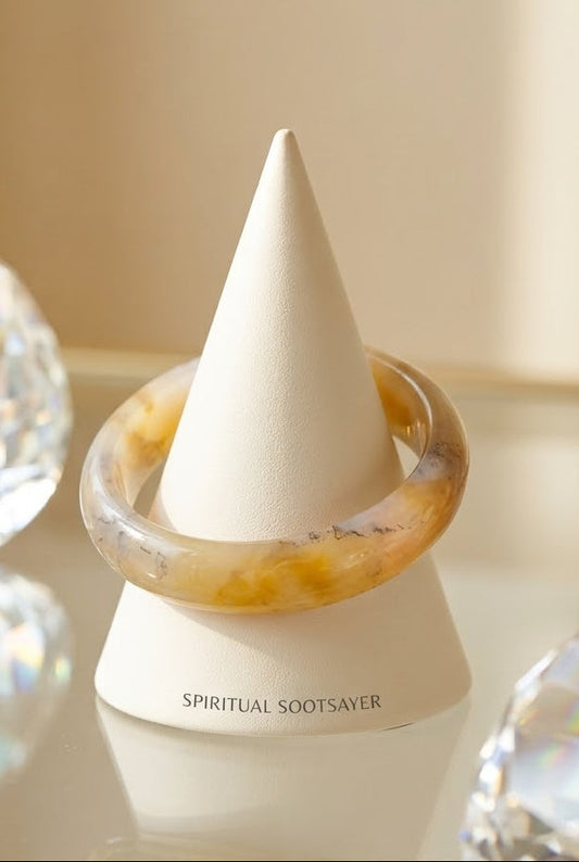 A translucent amber-yellow bangle bracelet displayed on a white cone stand labeled 'SPIRITUAL SOOTSAVER', with blurred faceted crystal-like props on both sides against a warm beige background.