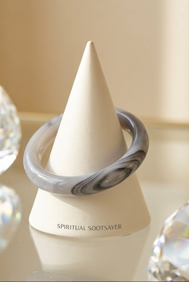 A gray and white marbled ring displayed on a cream cone stand, with the brand name 'SPIRITUAL SOOTSAVER' printed on the base. The stand is set on a reflective surface with blurred crystal spheres in the foreground and a soft beige background.