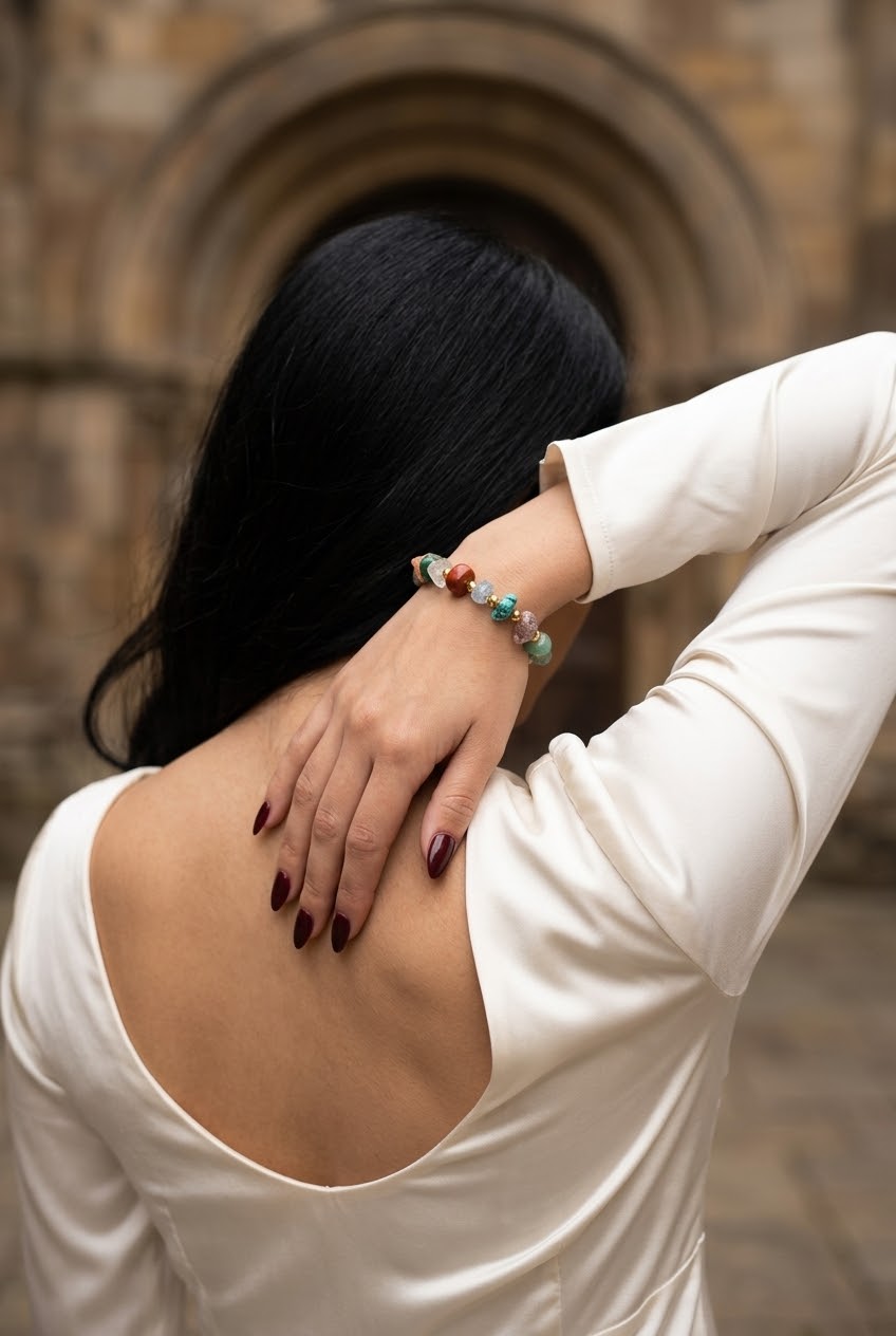 A woman with long black hair, seen from behind, wearing a cream-colored satin top and a colorful beaded bracelet on her wrist, posing in front of a stone archway.