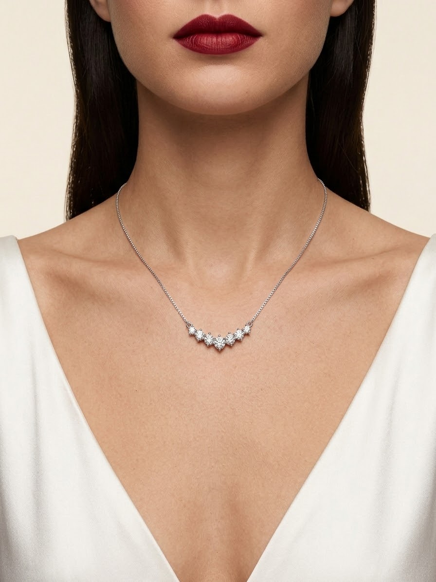 A close-up of a model wearing a delicate silver necklace with a curved row of small clear gemstones at the center, paired with a white V-neck top.
