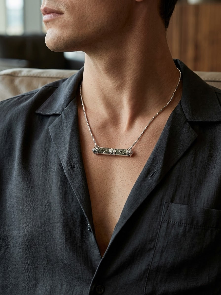 A close-up of a man wearing a silver bar necklace with textured green stones, styled with a dark gray linen shirt.