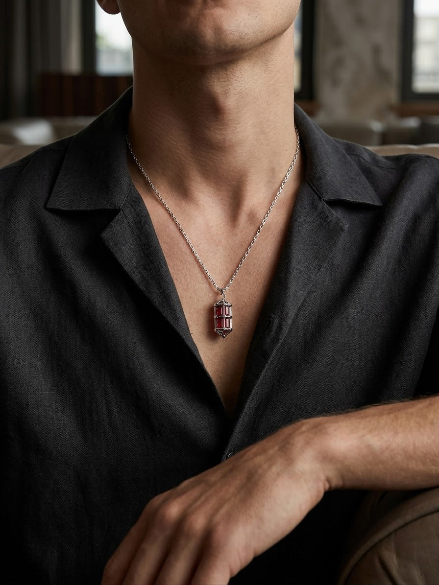 A close-up of a man wearing a silver-toned chain necklace with a small rectangular pendant featuring red and white striped details, styled with a dark gray linen shirt.