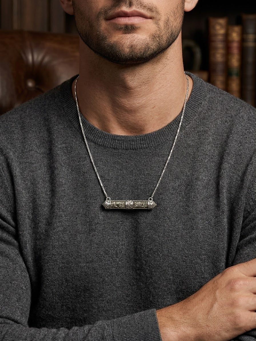 A man wearing a dark gray knit sweater and a silver-toned bar necklace with a textured, engraved horizontal bar pendant, set against a warm, blurred background of a leather chair and bookshelves.