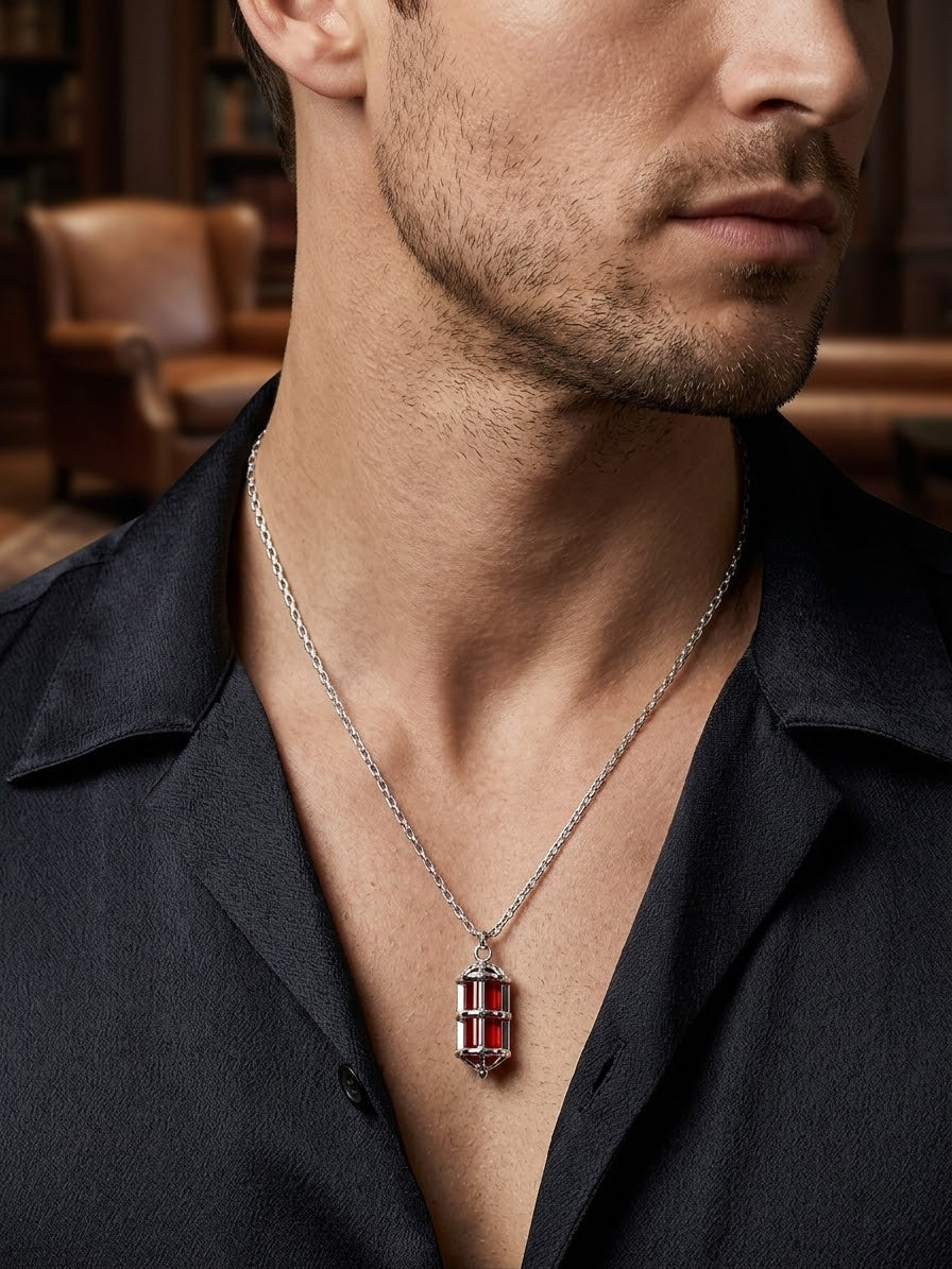 A close-up of a man wearing a silver-toned chain necklace with a rectangular pendant featuring red and black striped detailing, styled with a dark open-collar shirt in a warm, softly blurred indoor setting.