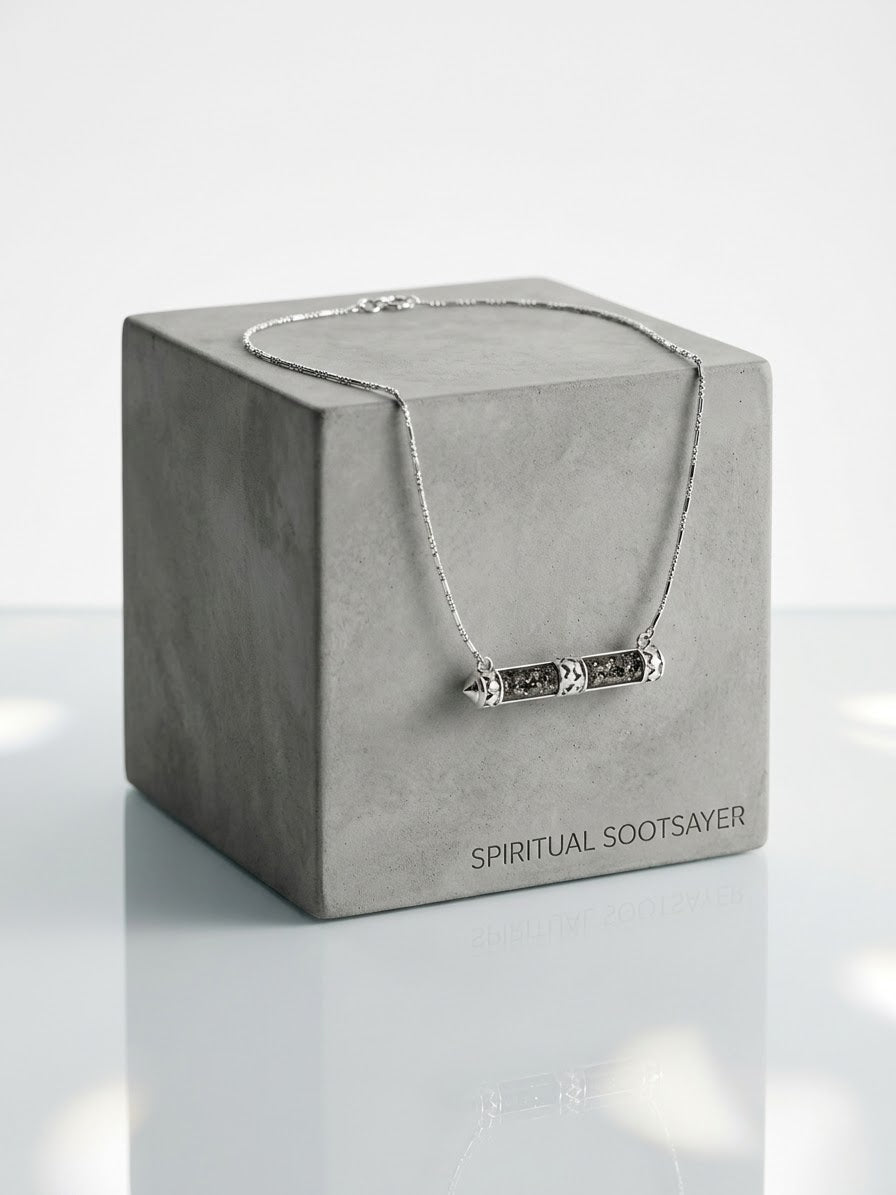 A silver-toned bar necklace with engraved details draped over a textured gray display box. The box is printed with the brand name 'SPIRITUAL SOOTSAVER' and is set against a clean, light gray background with a soft reflection below.