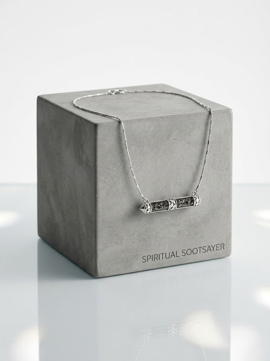 A silver-toned bar necklace with engraved details draped over a textured gray display box. The box is printed with the brand name 'SPIRITUAL SOOTSAVER' and is set against a clean, light gray background with a soft reflection below.