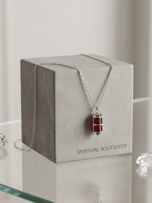 A silver-toned Spiritual Sootsayer necklace with a red cylindrical pendant, draped over a light gray display box on a reflective glass surface.