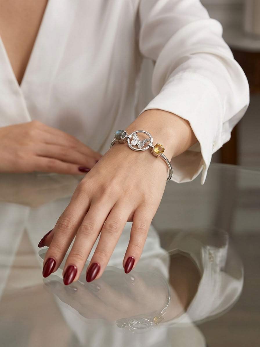 A close-up of a person's hand wearing a silver-toned bangle bracelet with a central circular charm and small gold and blue-green accents, styled with a white blouse and dark red nail polish.