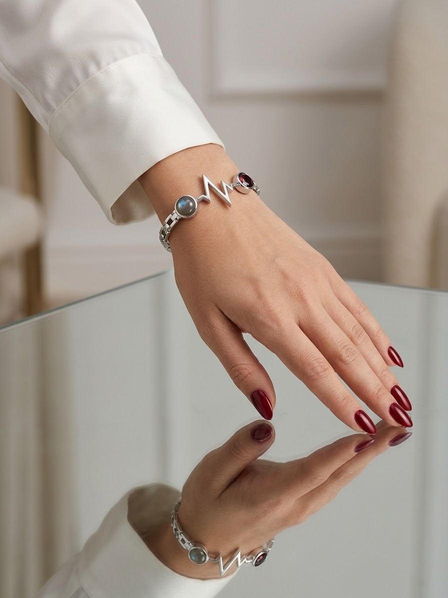 A close-up of a person's hand wearing a silver-toned bracelet with a lightning bolt charm and small round charms, paired with a white long-sleeve shirt and dark red nail polish, with a mirror reflection below.