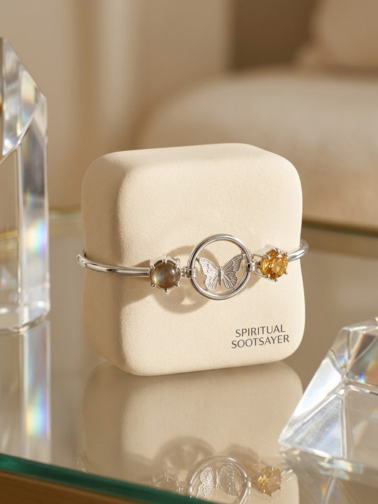 A silver-toned bangle bracelet with a butterfly motif and two small gemstone accents, displayed on a cream-colored jewelry box labeled 'SPIRITUAL SOOTSAYER'. The box is set on a reflective glass surface with blurred crystal and sofa elements in the background.