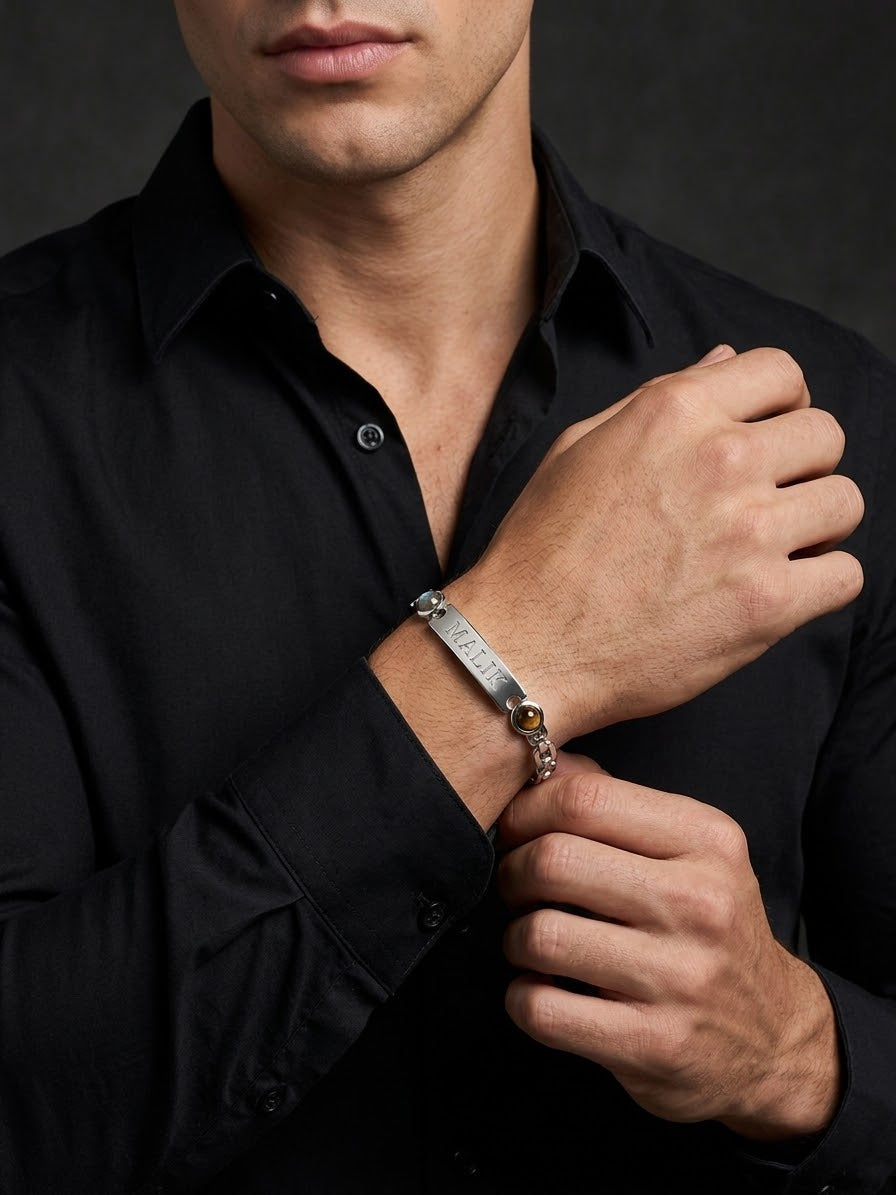 A close-up of a man wearing a silver engraved bracelet on his wrist, dressed in a black button-down shirt against a dark studio background.