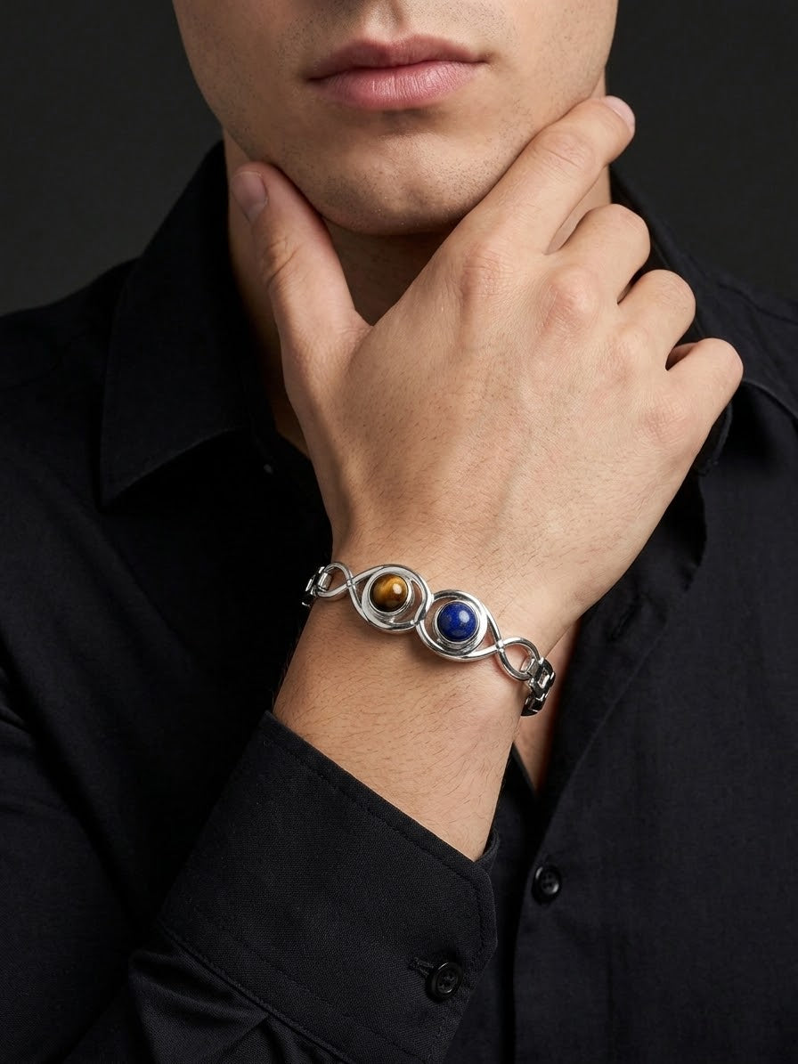 A close-up of a man wearing a silver-toned chain bracelet with three oval gemstone accents in brown, blue, and white, styled with a black shirt.