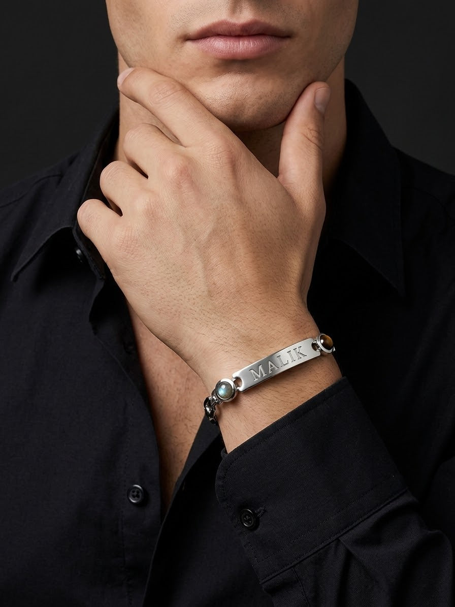 A close-up of a man wearing a silver bar bracelet engraved with the name 'MALK' and accented with small green stones, paired with a black button-down shirt.