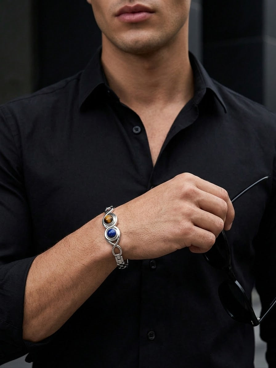 A close-up of a man wearing a silver-toned bracelet with a blue circular stone and a small amber accent, paired with a black button-down shirt.