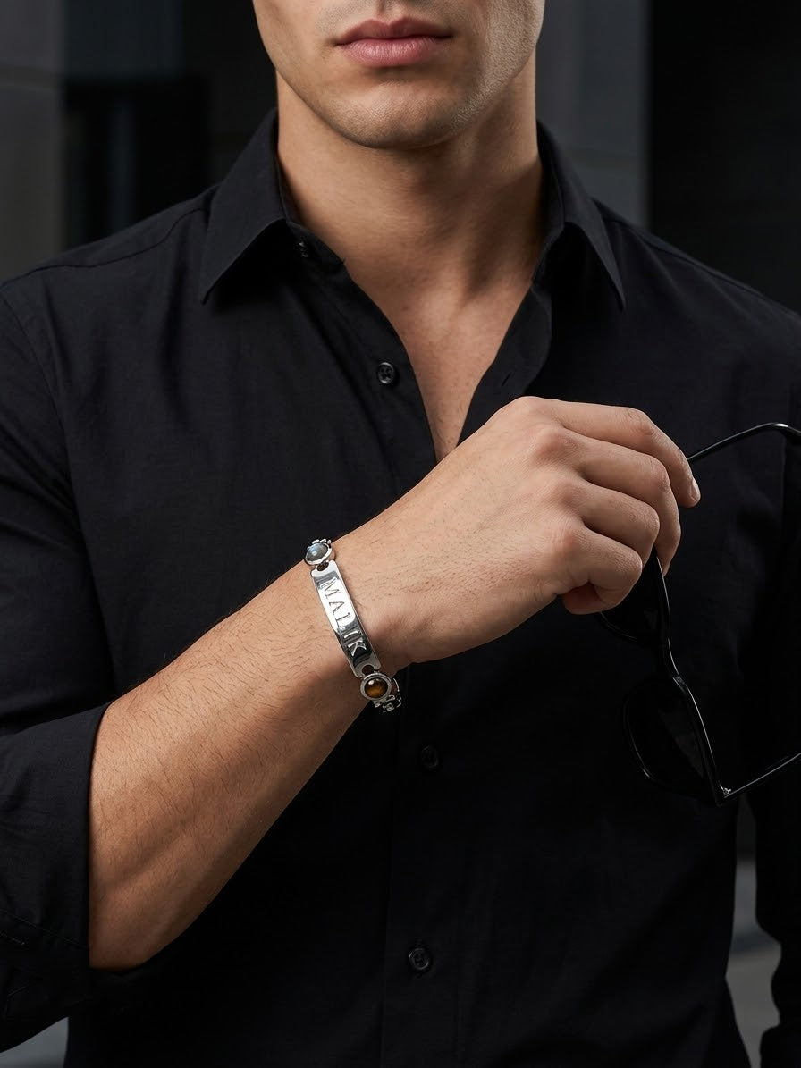A close-up of a man wearing a black button-down shirt and holding black sunglasses while wearing a silver-toned metal bracelet with a small amber stone on his wrist.