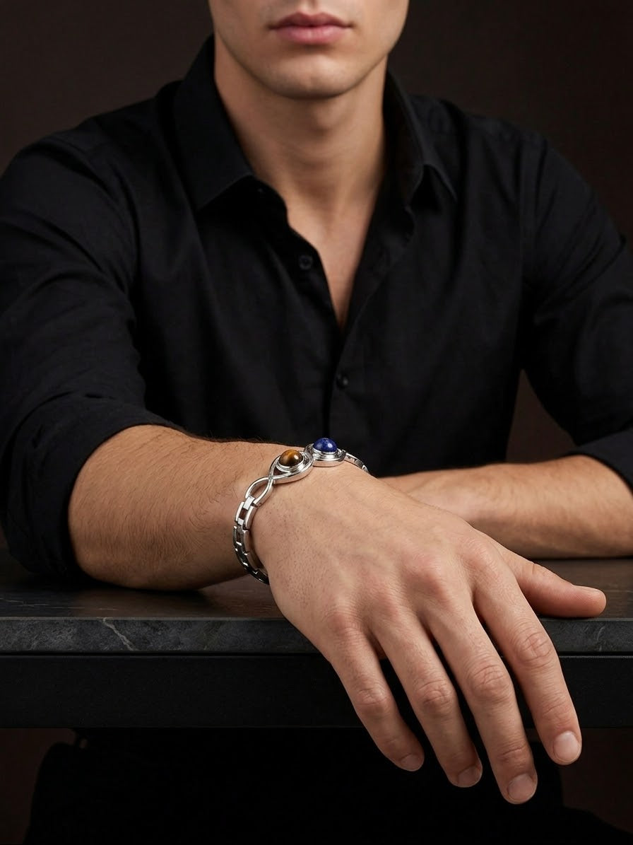 A man wearing a black button-down shirt and a silver-toned chain bracelet with a blue gemstone accent, resting his arm on a dark table.