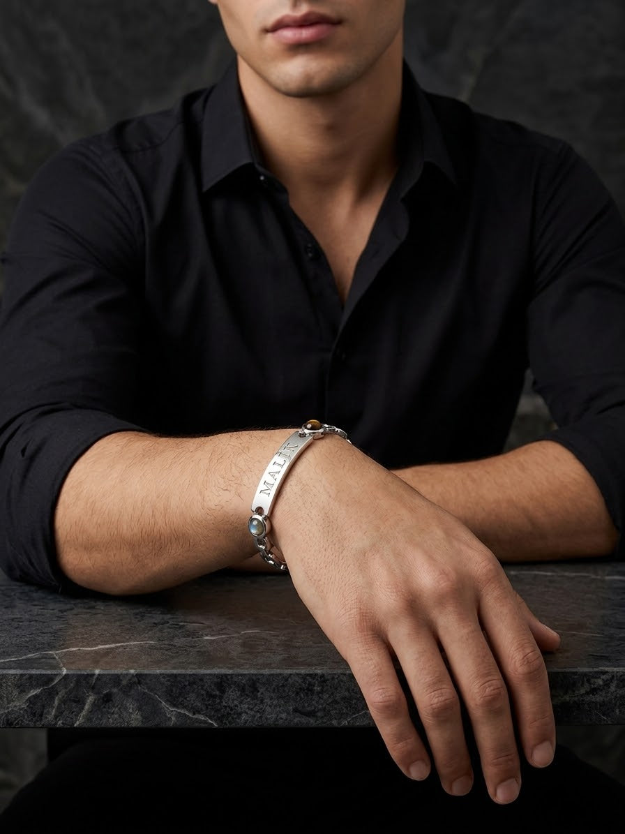 A man wearing a black button-down shirt and a silver-toned metal bracelet with engraved lettering, resting his arm on a dark marble surface.
