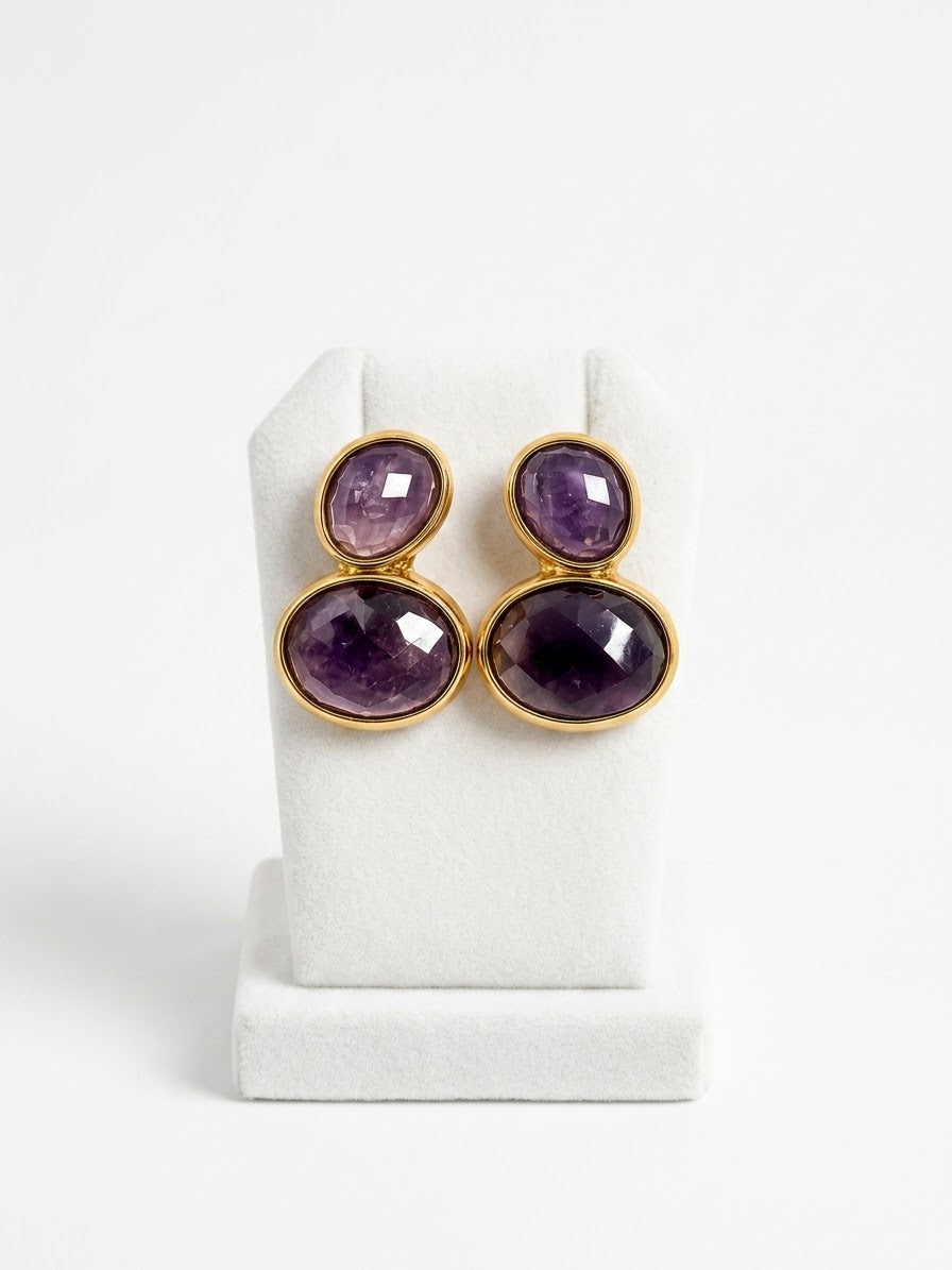 A pair of gold-tone drop earrings with oval purple amethyst gemstones, displayed on a white jewelry stand against a white background.