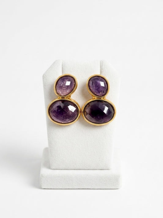 A pair of gold-tone drop earrings with oval purple amethyst gemstones, displayed on a white jewelry stand against a white background.