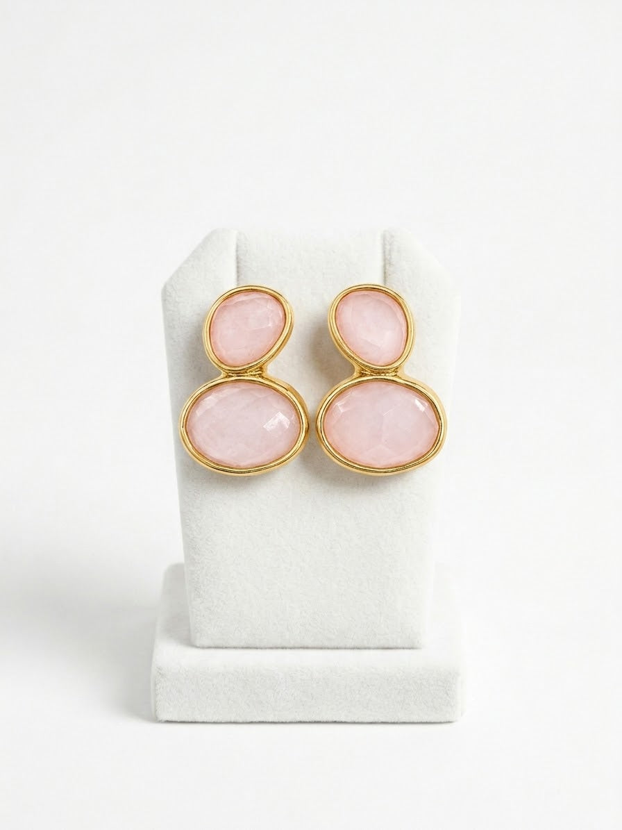 A pair of gold-tone drop earrings with oval pink gemstones, displayed on a white jewelry stand against a white background.