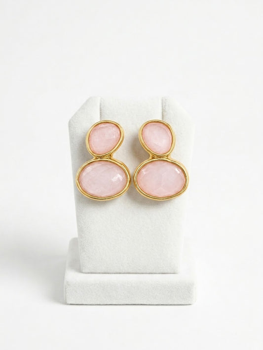 A pair of gold-tone drop earrings with oval pink gemstones, displayed on a white jewelry stand against a white background.