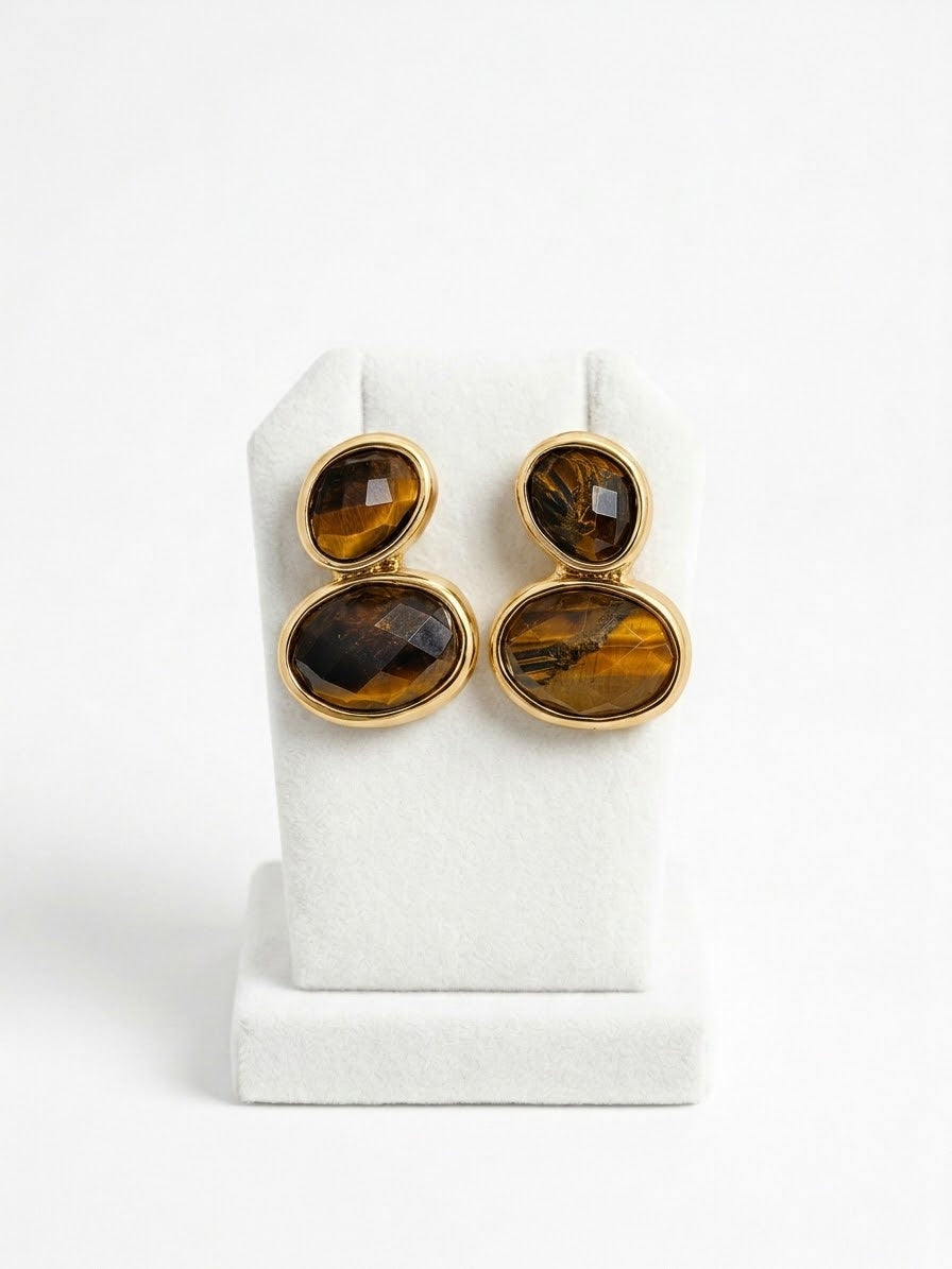 A pair of gold-toned drop earrings with stacked oval tiger's eye stones, displayed on a white jewelry stand against a white background.