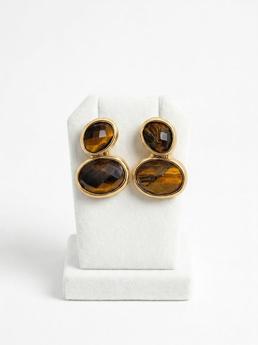 A pair of gold-toned drop earrings with stacked oval tiger's eye stones, displayed on a white jewelry stand against a white background.
