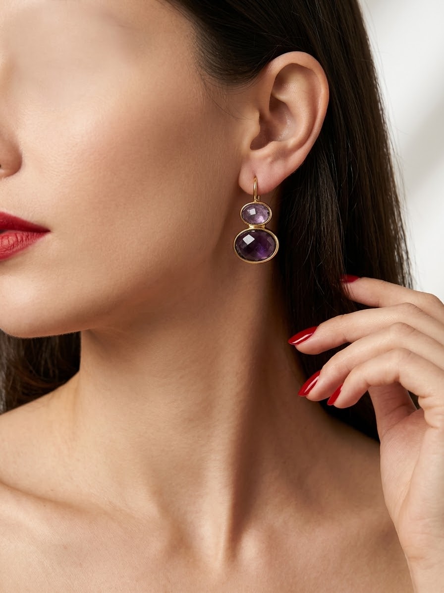 A close-up of a woman wearing a gold drop earring with two stacked oval purple gemstones, with her hand near her neck.