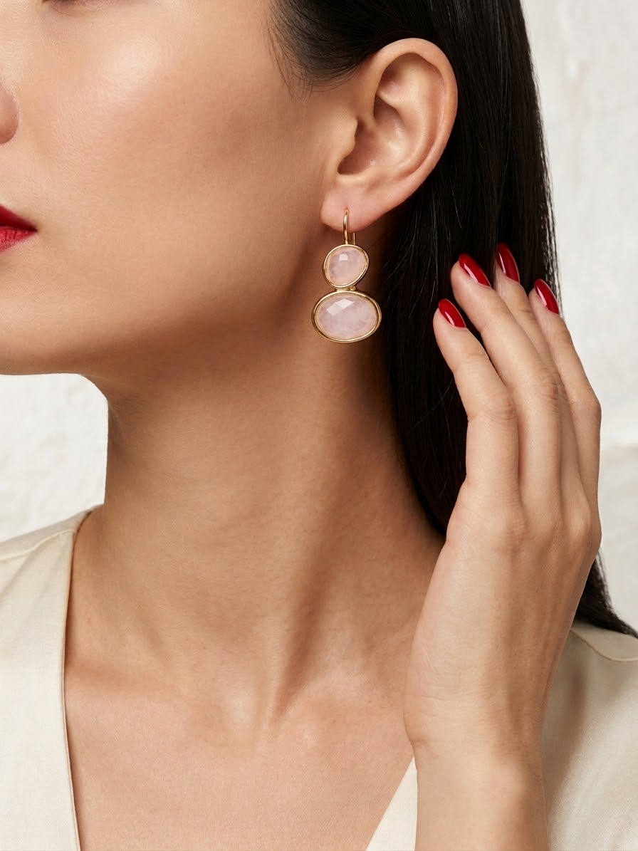 A close-up of a model wearing a gold drop earring with two pale pink stone charms, with her hand near her face.