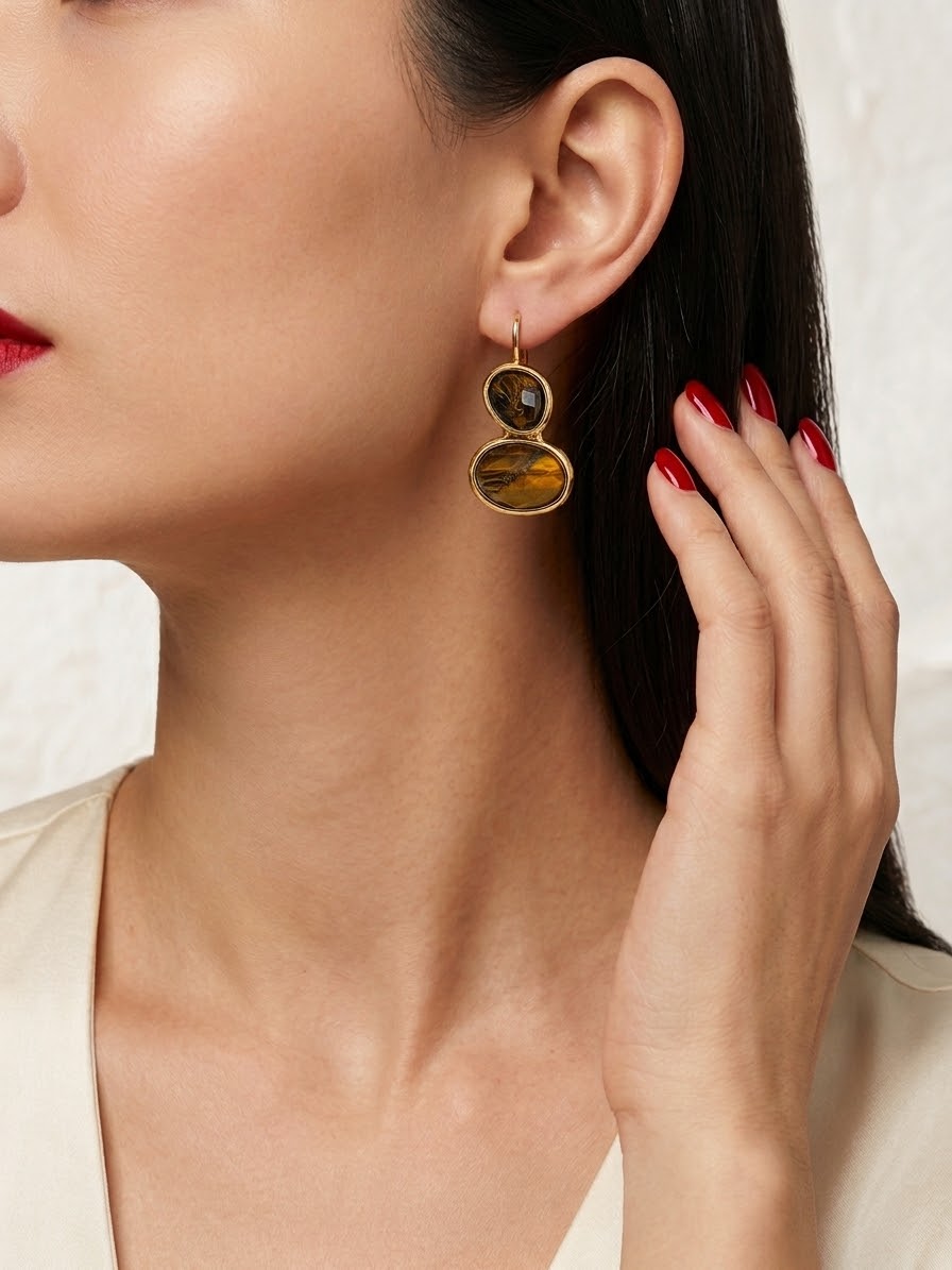 A close-up of a model wearing a gold dangle earring with two stacked oval tiger's eye stones, with her hand near her face.