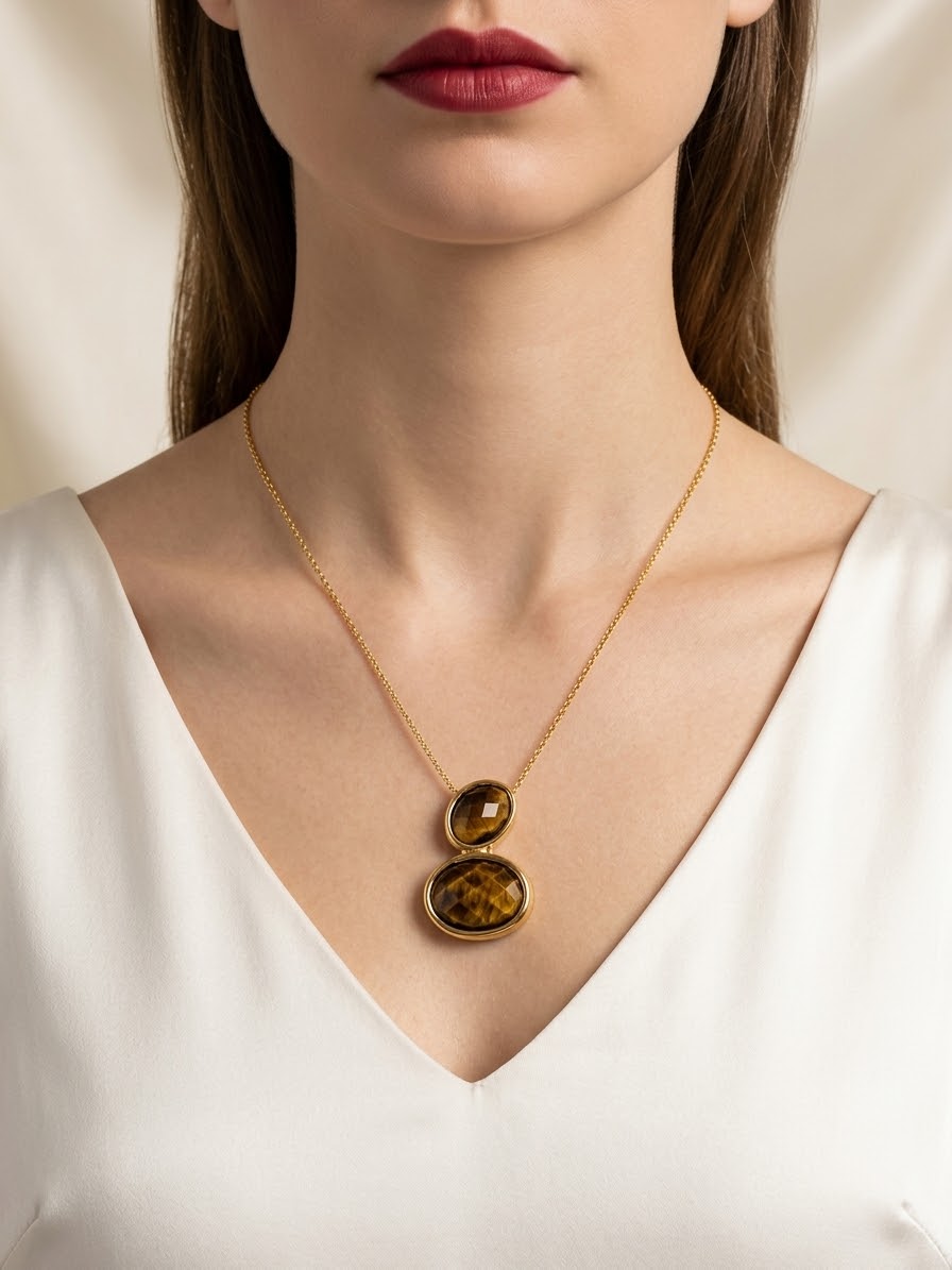 A close-up of a model wearing a delicate gold chain necklace with two stacked oval pendant charms featuring a textured golden-brown pattern, styled with a white V-neck top.
