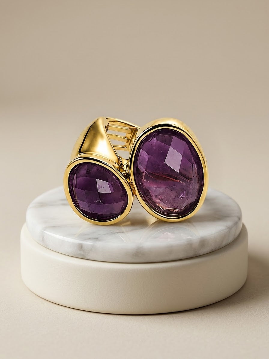 A gold ring with two large oval purple amethyst gemstones, displayed on a round white marble and cream pedestal against a warm beige background.
