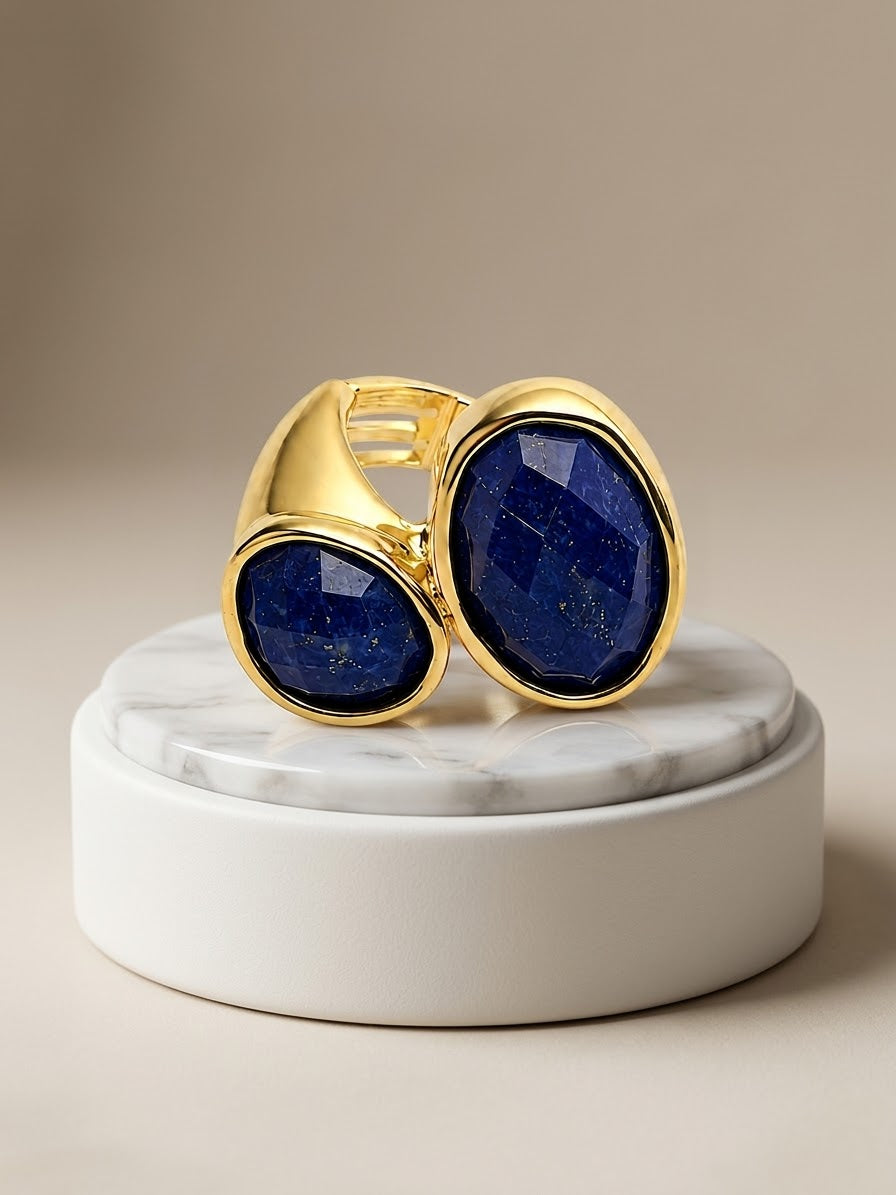 A gold ring with two large oval blue lapis lazuli stones, displayed on a white marble and white cylindrical pedestal against a warm beige background.