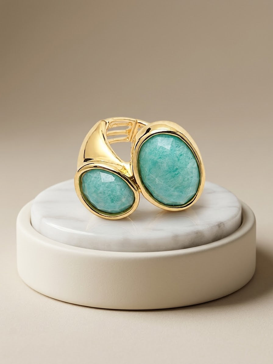 A gold-toned ring with two oval light aqua gemstones, displayed on a round white marble tray against a warm beige background.
