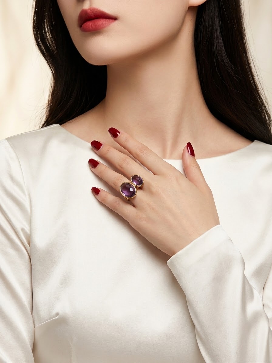 A close-up of a woman wearing a silver ring with two oval purple gemstones, resting her hand on her chest. She is wearing a white satin top and has dark red nail polish.