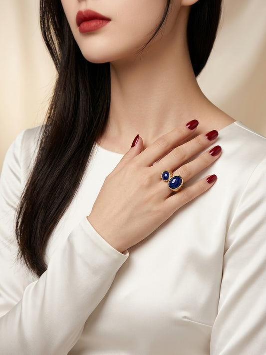 A close-up lifestyle shot of a model wearing a gold ring with two round blue gemstones, styled with dark red nail polish and a white satin top.