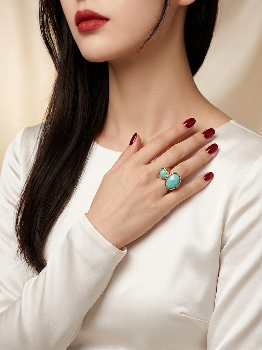 A close-up lifestyle shot of a model wearing a gold ring with two round blue gemstones, styled with dark red nail polish and a white satin top.