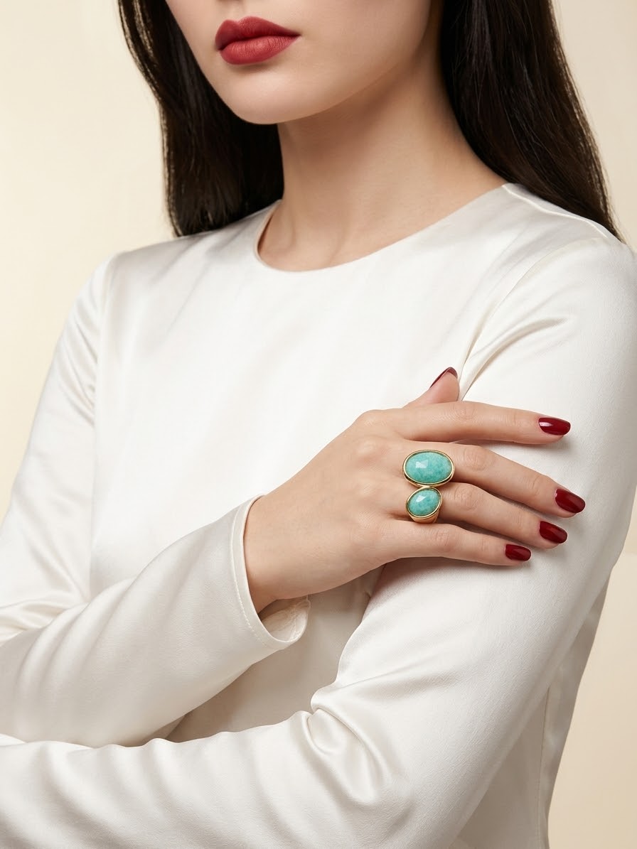 A close-up of a woman wearing a white long-sleeve top and a gold ring with two oval turquoise-colored stones, posed against a light beige background.