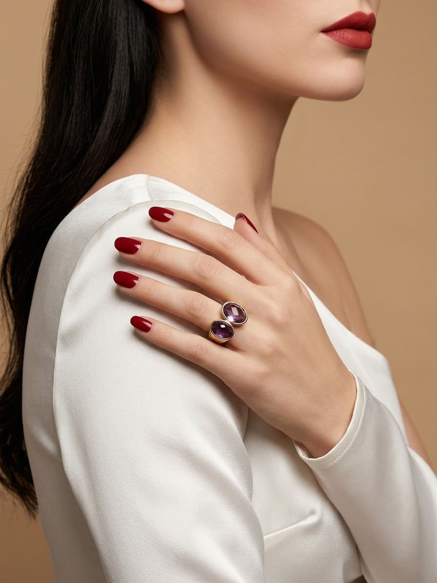 A close-up lifestyle shot of a model wearing a silver ring with two oval purple gemstones on her finger, paired with a white top and red nail polish against a warm beige background.