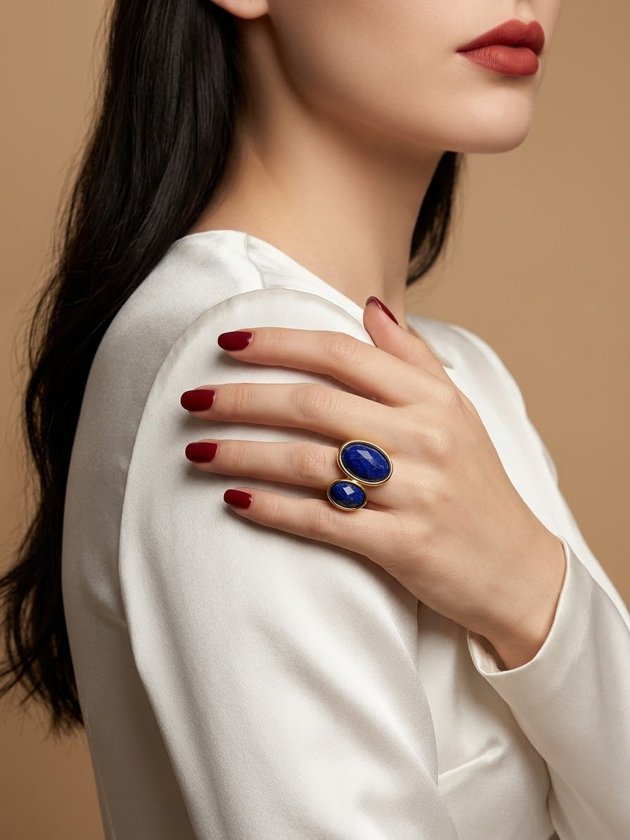 A close-up lifestyle shot of a model wearing a gold ring with two oval blue gemstones, set against a warm beige background.