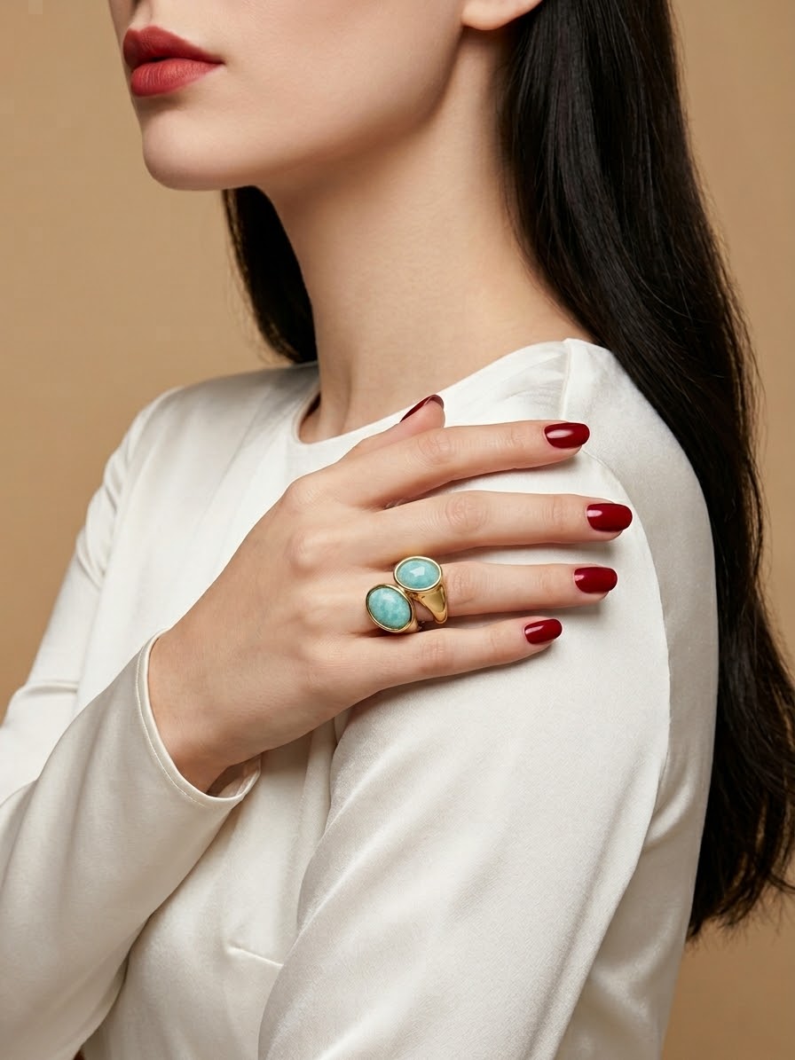 A close-up lifestyle shot of a model wearing a gold ring with two oval turquoise-blue stones, styled on her hand against a warm beige background.