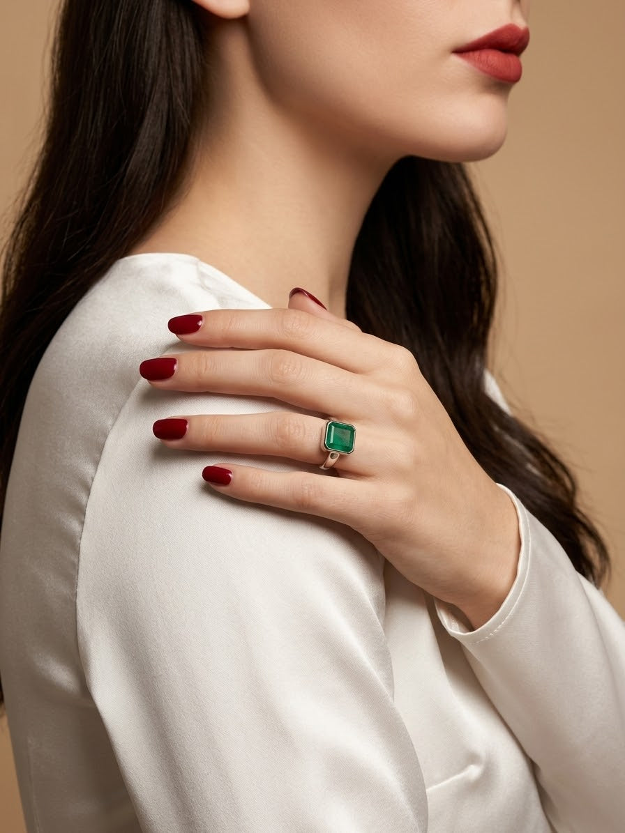 A close-up lifestyle shot of a model wearing a gold ring with a square-cut green emerald gemstone, styled on her finger with dark red nail polish against a warm beige background.