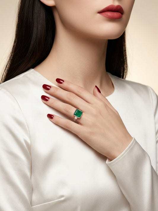 A close-up of a woman wearing a gold ring with a rectangular green emerald-cut gemstone, resting her hand on her chest. She is wearing a white satin top and has dark red nail polish.