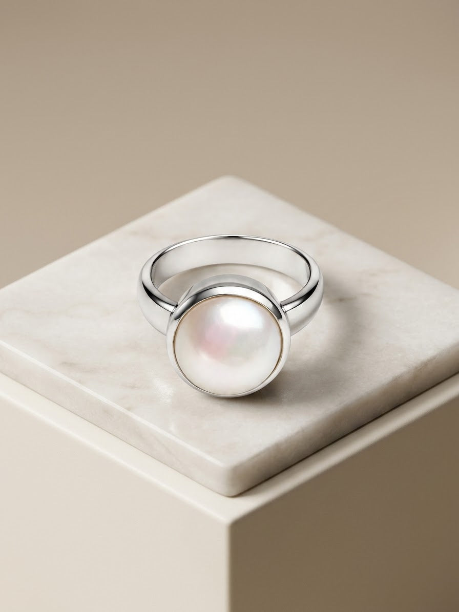 A silver ring with a large, round, lustrous white pearl bezel-set on a polished stone pedestal against a warm beige background.